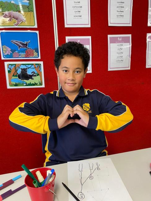 A student sits in front of his pencil drawing. He holds his hands at chest height in the shape of a heart.