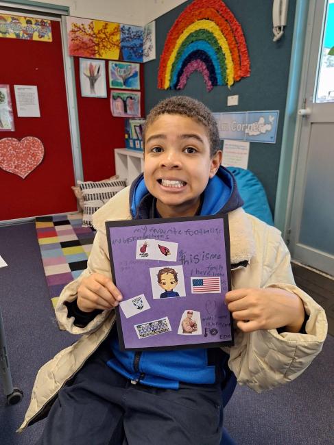 A student holds his completed collage. He is smiling. The collage has cut-outs of images on a deep purple background.