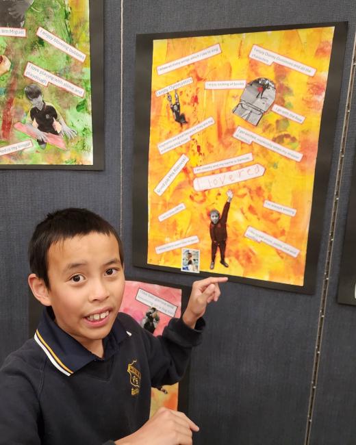 A student points at his collage hanging on the exhibition wall. It has black and white paper cut-outs of printed phrases and objects, including a picture of him on a bright yellow and orange background.