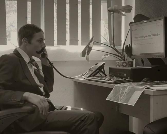 A still taken from the black and white film, ‘Charlie’s Crusade’. A student is dressed as the Deputy Principal, Mr Ismay. He wears a suit, a fake moustache and sits at a desk answering the phone.