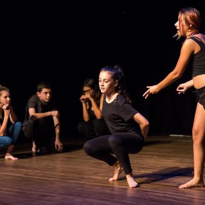 A performed scene with a student standing over another squatted student that has her arms behind her back. Other students are watching the scene from the edge of the performance space.