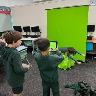 A boy using an iPad to film a scene in front of a green screen. Other students look on.