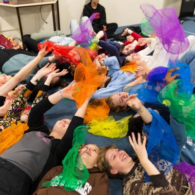 A group of students lie down with their heads together throwing different coloured scarves above them.